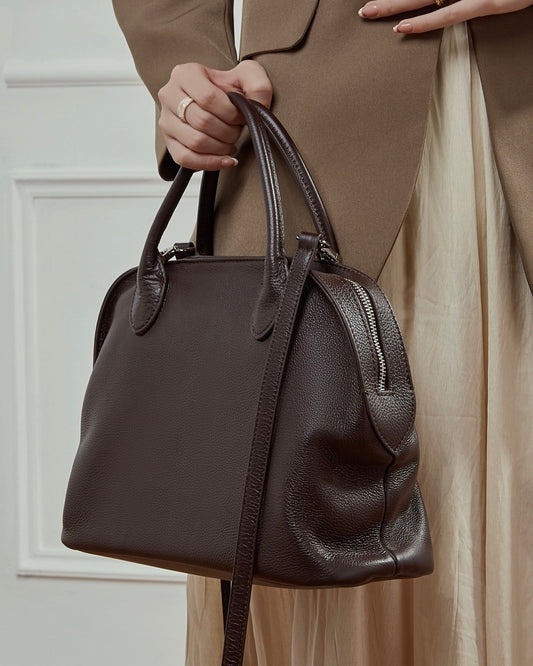 Brown leather handbag held by a person wearing a beige outfit.